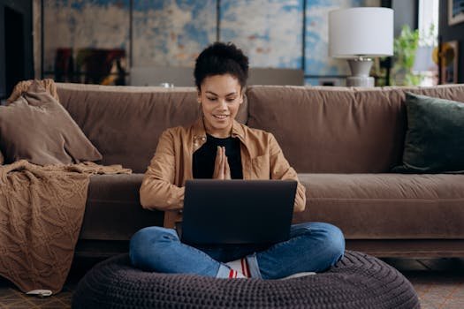 pexels-photo-6894013-6894013 A smiling young woman sits cross-legged on a pouf, using her laptop in a cozy home setting.